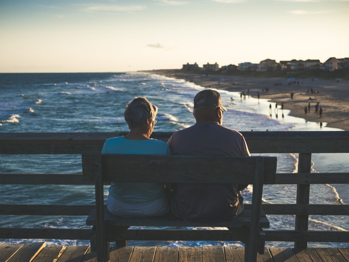 Elderly man and woman sit, facing the ocean on a pier during a sunny day.
