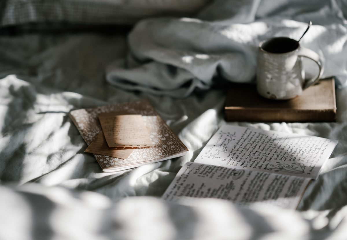 notebook with handwritten words is open on bed next to coffee cup and second notebook